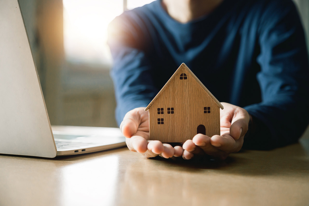 Hands holding a wooden model home with an open laptop