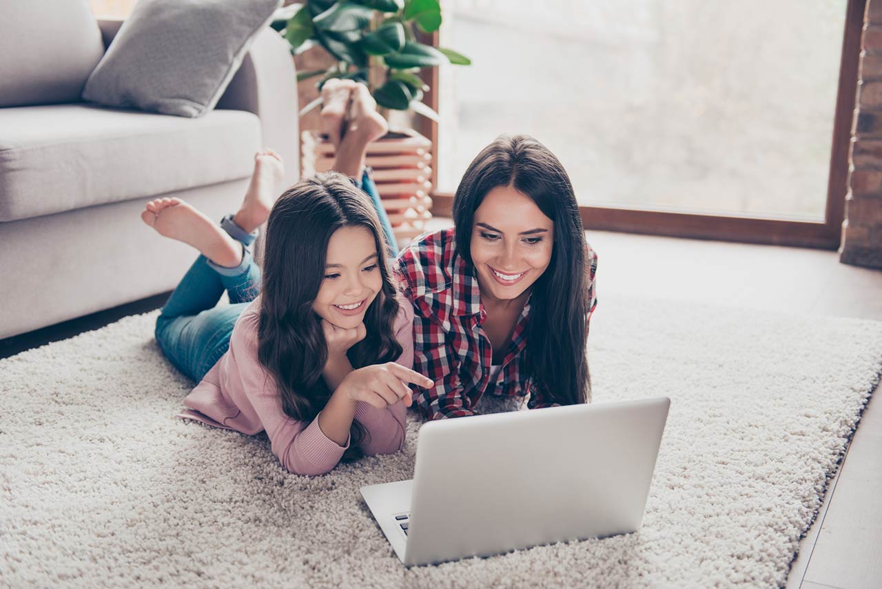 Mother and her little curious daughter are lying on a floor at home and using a laptop