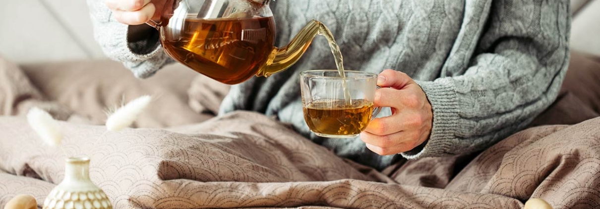 Woman dressed in knitted sweater pours hot tea from glass teapot into mug while sitting in bed