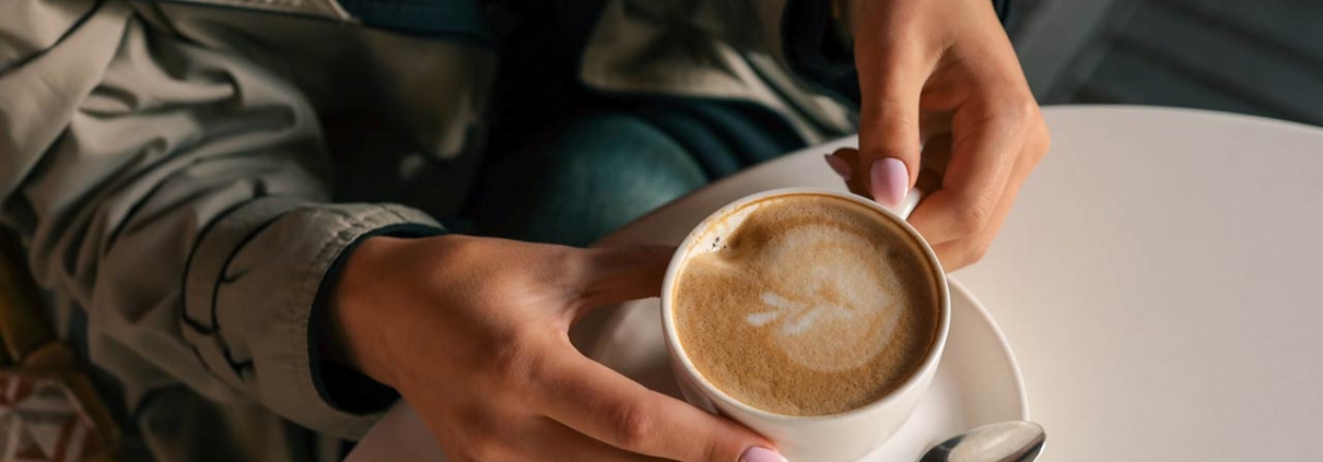 Close-up of woman's hands in trench coat gently holding a cup of cappuccino with latte art, seated at a white table
