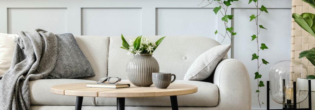 Living room interior with design grey sofa, wooden coffee table, tropical plants, shelf, mirror, furniture, plaid pillow, teapot, book and elegant personal accessories